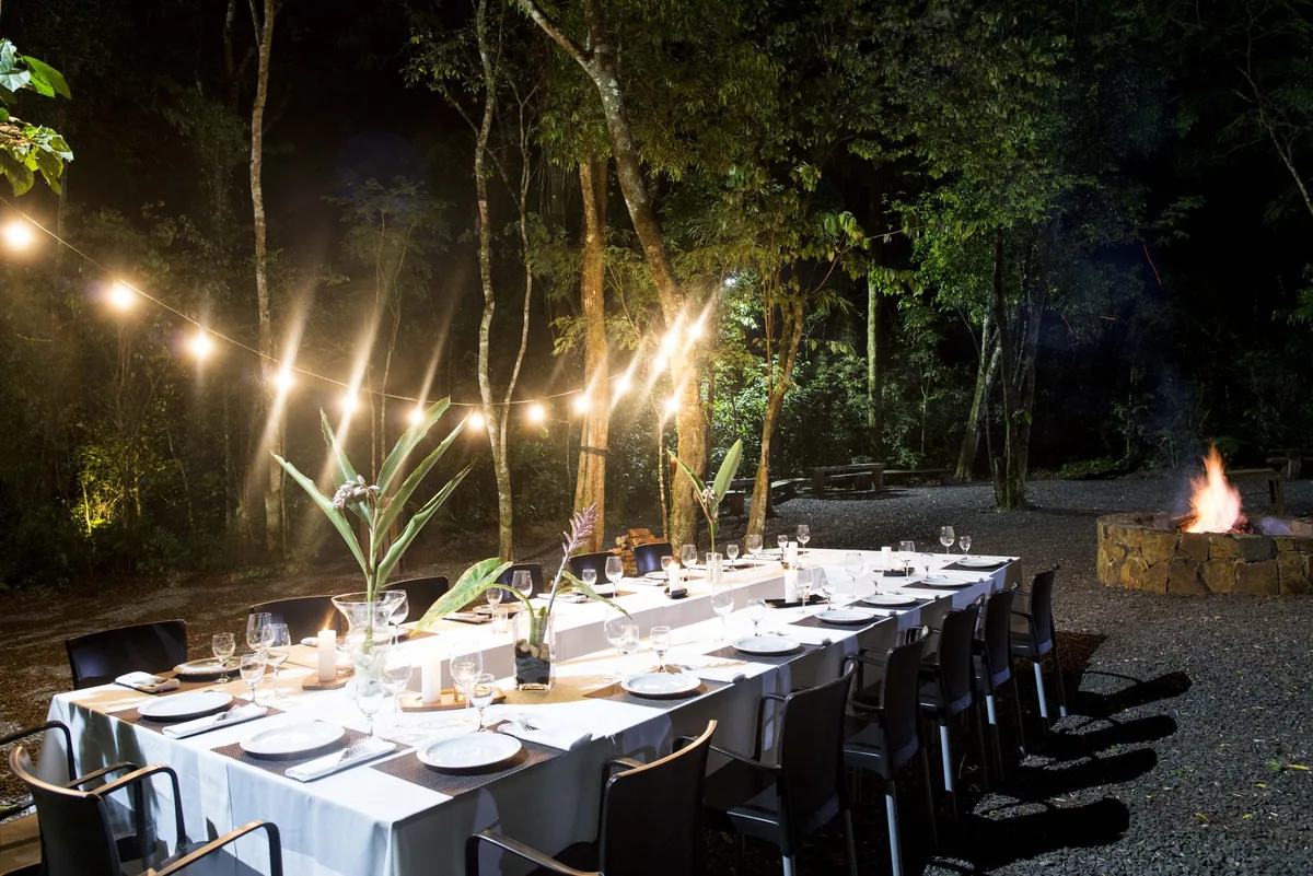 Mesa longa montada à noite no El Fogón, iluminada por varais de luz sob as árvores